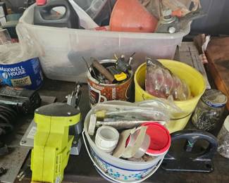 Tabletop collection of miscellaneous workshop items—cordless tools, yellow plastic tub filled with hardware, measuring tape, cords and shop supplies.