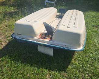 Two-person beige plastic pedal boat with two molded seats and foot pedals, sitting on a grassy lawn near water.