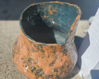 Small weathered copper pot with flared rim and handle, featuring green patina and rust, sitting outdoors on gravel.
