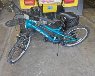 Youth-sized blue bicycle with black seat and handlebars, parked under a yellow ATV.