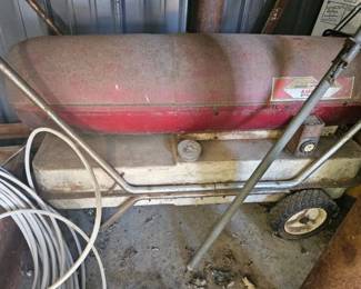 Red portable air compressor or fuel transfer tank with wheels and metal handle, attached hose coiled on top, sitting in the barn.