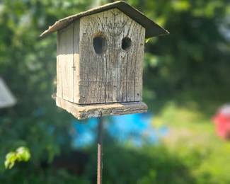 Rustic weathered wood birdhouse with pitched roof and small circular entry holes, hanging outdoors amid greenery.