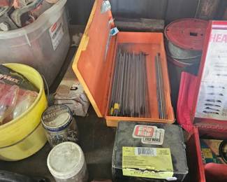 Assorted garage items including a car battery in a red plastic carrier, a yellow bucket with chain or rope, and a box of long metal rods.