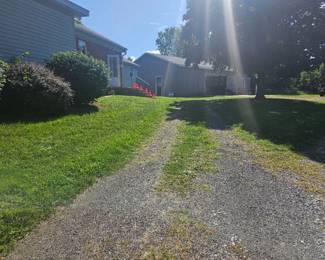 Gravel driveway leading from the front of the property, bordered by green grass and shrubbery under bright sunshine.