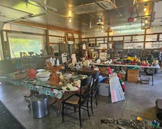 Large barn interior with long tables displaying assorted vintage items, tools, and collectibles under fluorescent lighting.