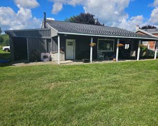Covered patio or carport attached to house with white posts and metal roof; large green lawn in front beneath blue sky with clouds.