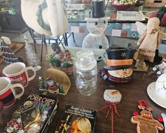 Assortment of holiday mugs, puzzles and decor on a table including Christmas-themed coffee mugs, decorative glass snowman jar, small snowman ornaments, star and tree figurines, and other festive knick-knacks.