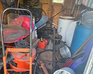 Barn interior packed with assorted equipment including an orange cement mixer on stand, red hand truck/dolly, blue barrel, buckets, hoses and long-handled tools.