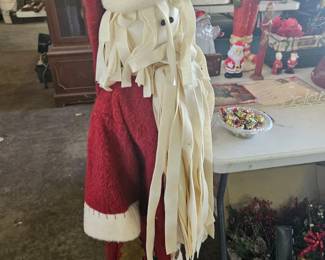 Tall Santa Claus or Saint Nick decorative figure wearing a red robe and long cream tassels, standing in front of tables filled with Christmas decor.