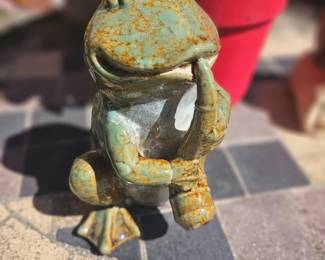 Charming cast stone or metal frog garden statue with a weathered green patina, sitting in a playful thoughtful pose on patio pavers.