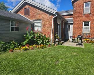 Rear courtyard view of brick farmhouse with colorful flower beds, green lawn and concrete walkway leading to a small patio seating area.