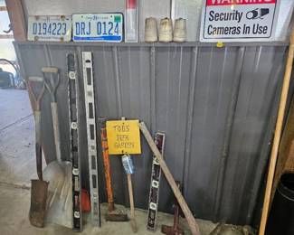 Collection of metal license plates, a security cameras sign, yellow yard sign and long metal stakes and poles leaning against the shed wall.