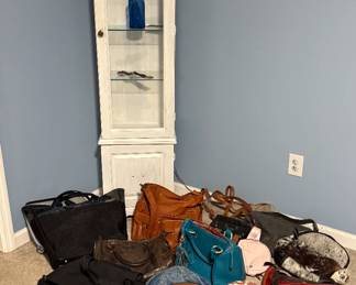 White Curio cabinet with glass shelves and light, various bags and purses including a couple of camera bags, a Myra cow print conceal carry backpack and Frye purse