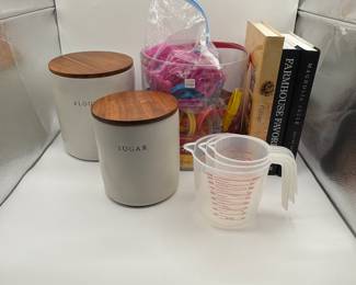 Two white storage containers with wooden lids, labeled 'SUGAR' and 'FLOUR'. A transparent container filled with colorful plastic cookie cutters. Three cookbooks with titles visible: 'College', 'Farmhouse Favorites', and 'Magnolia Table'. Several clear measuring cups with measurement markings. A metallic vented serving tray with handle. Also included is a large lot of cookie cutters.&nbsp;