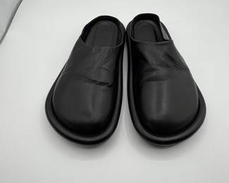 Black slip-on shoes featuring a smooth leather exterior, rounded toe shape, and a minimalistic design. The inside displays a soft lining, while the sole is slightly raised with a durable material. The brand name is embossed on the insole, showcasing a modern aesthetic.
