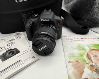 Includes a black Canon camera bag, a black Canon DSLR camera with a lens, a grey box for a Canon EF 75-300mm lens, various instruction manuals in color including a photo of a child and a cooking scene, and Canon branding on all items.