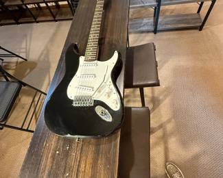 A black electric guitar with a glossy finish and a white pickguard. The body is shaped in a classic style. The headstock is natural wood with the text 'Starcaster' and 'Fender' branded logo. The fretboard is dark brown with metal fret markers. Accompanying the guitar is a black gig bag with the logo 'Epiphone' displayed on it.
