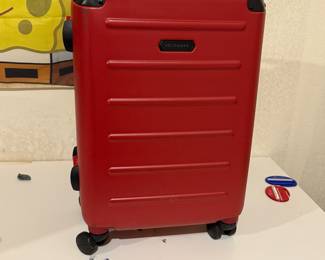 A bright red hard-shell suitcase featuring a vertical ribbed design. It has a black handle on top, four spinner wheels for easy maneuverability, and a TSA-approved combination lock. The front bears a small hexagonal logo, and the overall construction appears durable.