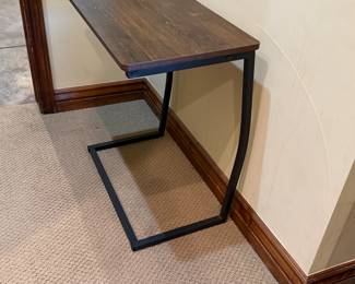 Features a rectangular wooden top in a dark brown finish, supported by a sleek black metal frame. The design is minimalist with a unique C-shape base providing stability. The dimensions are approximately 3 feet tall and 2 feet wide.