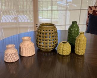 A collection of ceramic vases in various shapes and sizes. Includes a cream-colored textured vase, a green vase with a leaf design, a small light green vase with a similar leaf pattern, and a larger olive green woven lantern. The lantern features an open weave design and a round shape, while the vases have a glossy finish and intricate detailing.