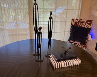 Three black metal stick figure sculptures of varying heights, with a tall slender figure, a medium figure, and a small figure. The sculptures are placed on a dark wooden round table. There are also two stacked books, one black with white stripes on the cover, and another white with black stripes on the cover. Atop the books, there is a black "jack" sculpture. The table features a geometric wood pattern.