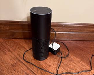A black cylindrical smart speaker with a mesh pattern on the lower half. It features a flat top with circular controls. The base has a logo that reads 'amazon'. A power adapter is attached with a cable coming out from the bottom