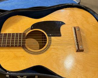 An acoustic guitar with a natural wood finish and a round sound hole. The guitar features a black pickguard and a wooden bridge. The headstock is mahogany with six tuning pegs, three of which are white. The guitar comes in a black hard case with a blue interior lining, shaped to fit the guitar snugly. The case has metal latches and a textured exterior.