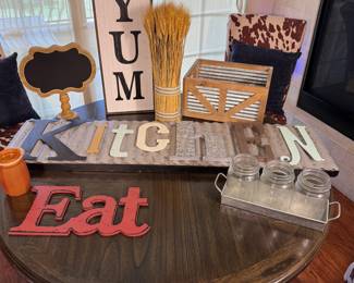The set includes a large distressed wooden sign with the word 'Kitchen' in multi-colored letters, a red decorative sign reading 'Eat', a tall vertical sign with the word 'YUM' in black text, a rustic wooden crate, an orange ceramic container, a small black chalkboard sign, and a tray containing three glass mason jars