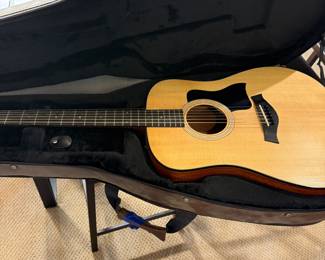 An acoustic guitar with a natural wood finish, featuring a black pickguard and six strings. The guitar is identifiable by the brand name 'Taylor' on the headstock. Accompanied by two guitar cases: a vintage brown hardshell case and a the original tan soft case, each with zippers and straps