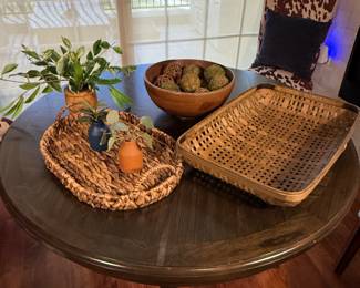 Set includes a large rectangular woven basket in brown with a squared pattern, a circular wooden bowl in a natural wood finish containing decorative orbs in green and brown hues, a small oval woven tray in light brown, and two ceramic planter pots—one in terracotta with a small green plant and another in navy blue with a leafy plant.
