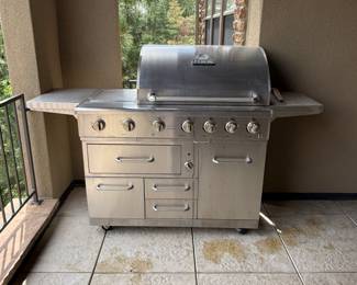 This stainless steel propane grill features a sleek silver finish, a rounded hood, six control knobs, and multiple storage drawers and compartments. It includes a side burner with a black grate, a cooking surface with metal grates, and a propane tank compartment. The brand logo 'NXR' is visible on the grill's hood. The grill has two stainless steel food pans and various grilling tools visible in the drawers.

Overall lengths is 6 feet overall depth, approximately 26 inches overall height, approximately 49 inches