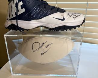 A blue and white Nike football cleat the Autograph of Dalton Risner #66. The cleat has black studs on the sole and a logo on the side. Accompanying the cleat is a white football, also signed, displayed in a clear acrylic case.