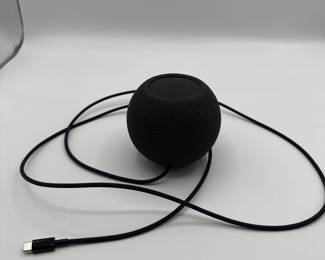 A spherical black smart speaker with a textured surface. The top features an orange touch control panel with plus and minus symbols. The bottom displays the Apple logo. It comes with a black USB-C charging cable.