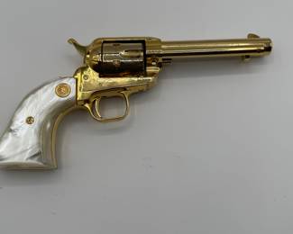 A gold-plated .22 Colt Collectible revolver featuring a pearl-colored grip. The gun has a shiny, metallic finish with a distinct shape, showcasing ornate detailing. Engraved text on the barrel reads '1864-1964 KANSAS SERIES WICHITA-1984.' The grip has a gold emblem and the overall design is a classic revolver style.