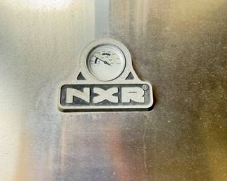 This stainless steel propane grill features a sleek silver finish, a rounded hood, six control knobs, and multiple storage drawers and compartments. It includes a side burner with a black grate, a cooking surface with metal grates, and a propane tank compartment. The brand logo 'NXR' is visible on the grill's hood. The grill has two stainless steel food pans and various grilling tools visible in the drawers.

Overall lengths is 6 feet overall depth, approximately 26 inches overall height, approximately 49 inches