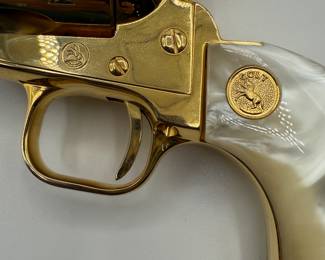 A gold-plated .22 Colt Collectible revolver featuring a pearl-colored grip. The gun has a shiny, metallic finish with a distinct shape, showcasing ornate detailing. Engraved text on the barrel reads '1864-1964 KANSAS SERIES WICHITA-1984.' The grip has a gold emblem and the overall design is a classic revolver style.