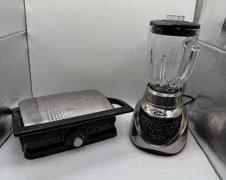 Set includes a stainless steel blender with a glass pitcher and a grilled sandwich maker with a stainless steel lid. The blender features a silver base with black buttons. The sandwich maker has a flat cooking surface with black ridges and a control knob with red and green indicator lights.