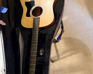 An acoustic guitar with a natural wood finish, featuring a black pickguard and six strings. The guitar is identifiable by the brand name 'Taylor' on the headstock. Accompanied by two guitar cases: a vintage brown hardshell case and a the original tan soft case, each with zippers and straps