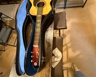 An acoustic guitar with a natural wood finish and a round sound hole. The guitar features a black pickguard and a wooden bridge. The headstock is mahogany with six tuning pegs, three of which are white. The guitar comes in a black hard case with a blue interior lining, shaped to fit the guitar snugly. The case has metal latches and a textured exterior.
