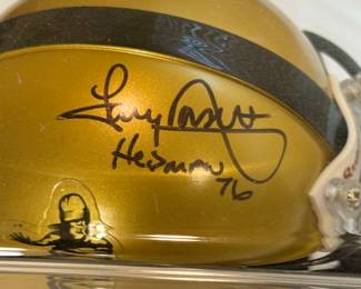 A gold mini helmet with a black stripe, featuring the Heisman Trophy logo on one side and autographed by Tony Dorsett. The helmet has an autograph by a Heisman winner on the top, along with the inscription 'Heisman 76'. The helmet is displayed in a clear acrylic case on a black pedestal with gold accents, which includes an engraved plate that reads 'HEISMAN TROPHY FRESHMAN OF THE YEAR TONY DORSETT.”