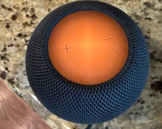 A spherical black smart speaker with a textured surface. The top features an orange touch control panel with plus and minus symbols. The bottom displays the Apple logo. It comes with a black USB-C charging cable.