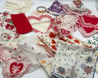 A collection of vintage handkerchiefs featuring various colors and patterns. Includes red, white, purple, and floral designs. Some handkerchiefs have heart shapes and embroidered flowers. A gold-tone bracelet watch is also visible, alongside intricately designed lace edges and colorful floral motifs.