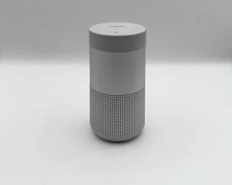 Gray portable smart speaker with a cylindrical shape. Features a perforated speaker grille at the bottom. Top control panel includes buttons for power, Bluetooth, volume, and additional controls. The brand name 'BOSE' is prominently displayed on the top surface.