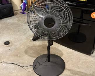 Black stand fan with a round base, metal blades, and a circular grille. Features the brand name 'Lasko' in the center. Adjustable height with a measuring tape showing the size.