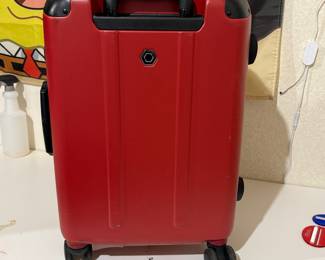 A bright red hard-shell suitcase featuring a vertical ribbed design. It has a black handle on top, four spinner wheels for easy maneuverability, and a TSA-approved combination lock. The front bears a small hexagonal logo, and the overall construction appears durable.