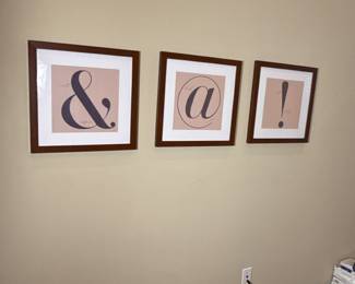 A set of four framed prints showcasing typography symbols. The first print features an ampersand '&' on a light beige background with a dark grey design. The second print displays the '@' symbol in a circular design, also in dark grey, set against the same light beige background. The third print contains an exclamation mark '!' presented in dark grey on a light beige backdrop, all framed in wooden frames with a brown finish.