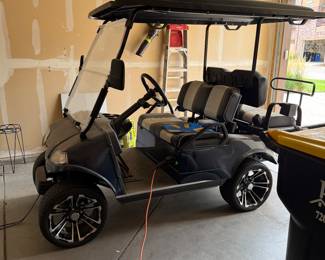 
EVolution Black electric golf cart with a canopy top, featuring two-tone seating in black and gray. The cart has custom black wheels with a stylish design, a steering wheel with a silver center logo, and various dashboard controls. The exterior includes a clear windshield and headlights, with visible wiring inside. Powers on and appears to be in good working order. See photos for front end damage. 