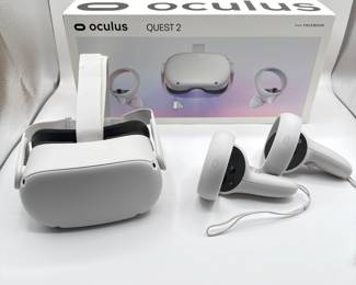 The item features a white VR headset with a sleek design, showing a curved front and side panels. It has a comfortable black foam cushion inside. The controllers are also in a white color, with buttons and an analog stick visible. The packaging includes the brand name 'Oculus' and the model 'Quest 2', along with a colorful graphic background.