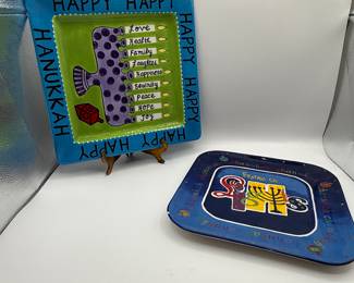 Two decorative plates featuring vibrant colors. The first plate is square with a bright blue border, green center, and white text spelling 'HAPPY HANUKKAH' around the edges. It features a purple menorah design and listings of words like 'Love', 'Health', and 'Joy' in black ink. The second plate is square with a dark blue background adorned with colorful text that reads 'Festival of Lights' and includes a menorah symbol in the center. Both plates are made of ceramic and display playful, holiday-themed artwork.