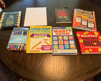 Contains a box of 72 watercolor pencils in a blue package, a clear bag of dual tip markers featuring various colors, a large print word-find book labeled 'Jumbo', a puzzle book titled 'Variety Puzzles', a 'True Crime Puzzle Box' book, a large print word search book titled '9000 Words', and a 'BuzzFeed Ultimate Book of Quizzes'. There is also a plain white sheet of paper.